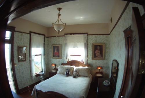 A cozy bedroom with floral wallpaper, a large bed, vintage paintings, and soft lighting from a chandelier.